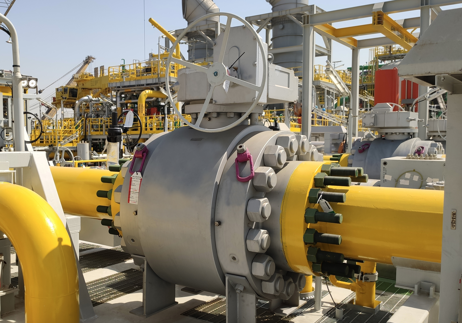 A large industrial facility with intricate machinery and yellow pipelines. The centerpiece is a massive valve with a circular handwheel, flanked by other equipment and towers in the background. The sky is clear and the overall setting appears to be a gas or oil processing plant.