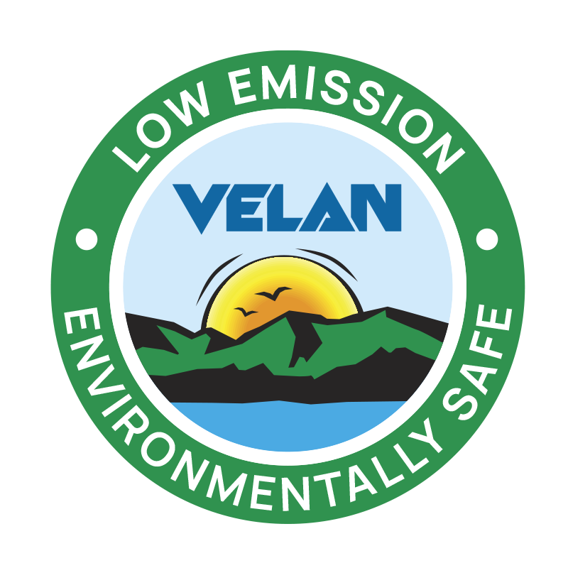 Green circular logo with the text "LOW EMISSION" at the top and "ENVIRONMENTALLY SAFE" at the bottom. Inside, a sunset scene with mountains and a lake is depicted, with the word "VELAN" above the image.