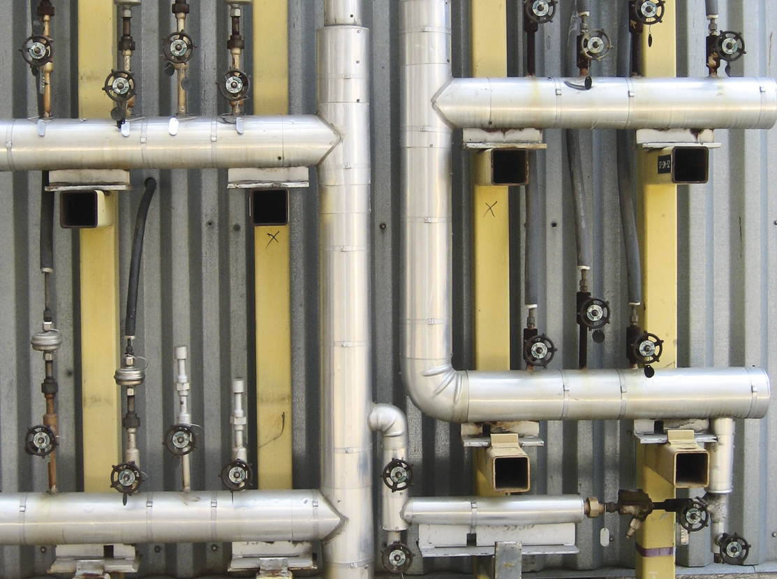 Pipes and valves with metal coverings are attached to a corrugated metallic wall, arranged vertically and horizontally. The setup includes several small hand-turn valves and features various shades of silver and yellow.