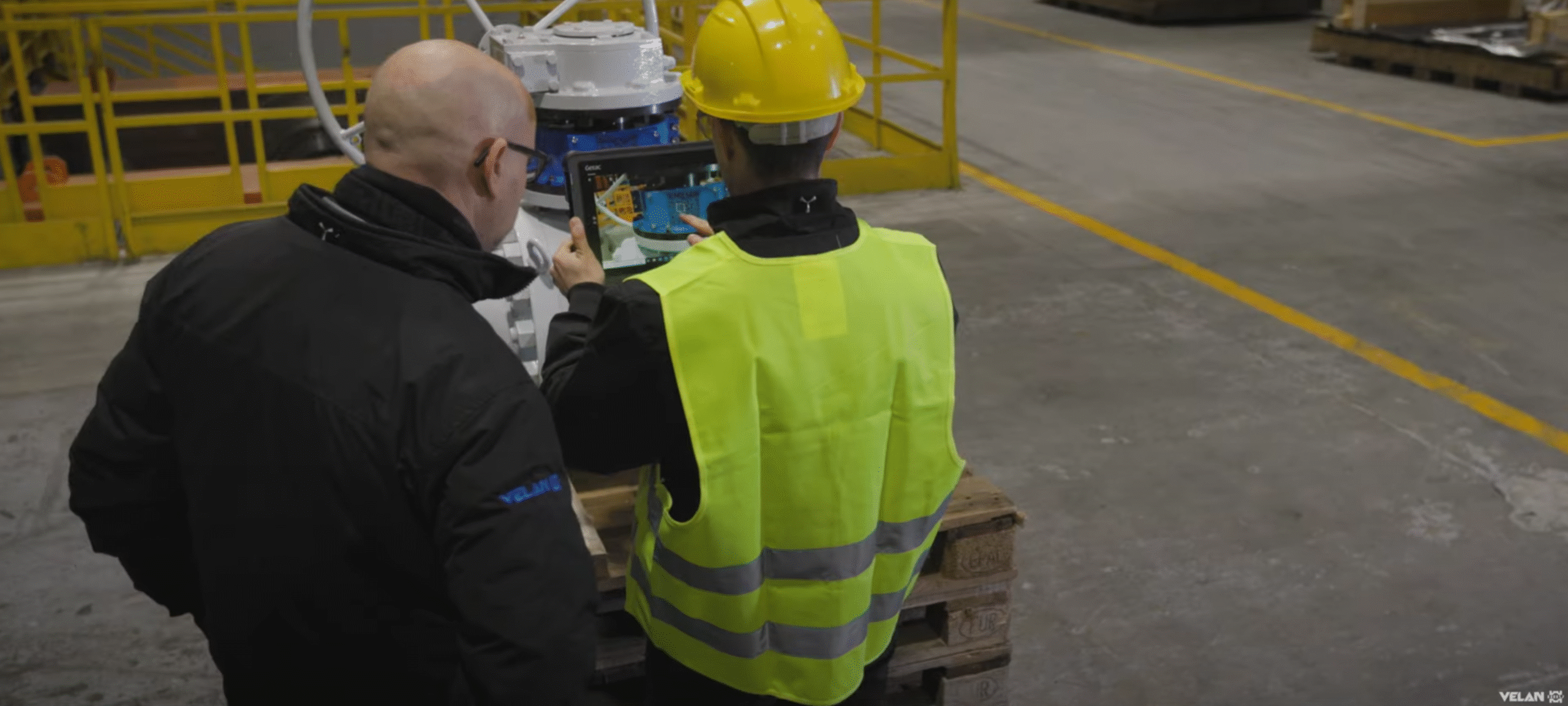 Deux travailleurs en tenue de sécurité, dont l'un porte un casque jaune et un gilet de haute visibilité, se tiennent dans un entrepôt pour examiner des équipements et utiliser une tablette.