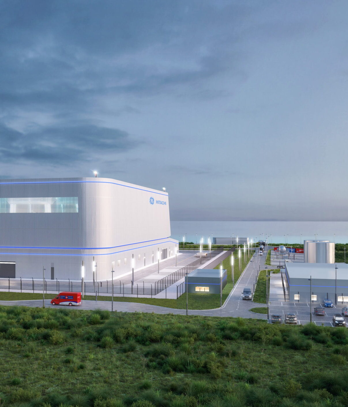 Un bâtiment industriel moderne et blanc avec des accents bleus est entouré d'une clôture et de verdure sous un ciel nuageux, avec des voitures garées le long des routes adjacentes et des structures plus petites à proximité.