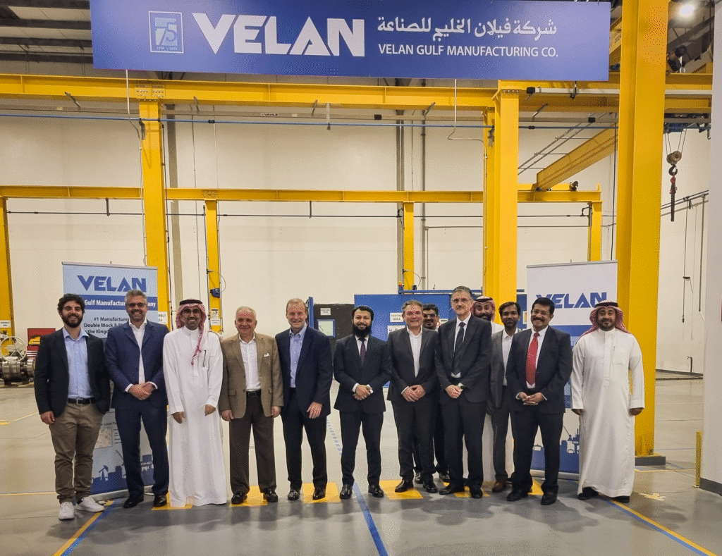 A group of thirteen men in business attire and traditional Middle Eastern clothing stand smiling under a large Velan Gulf Manufacturing Co. sign in a yellow industrial facility. Two Velan banners are visible in the background.