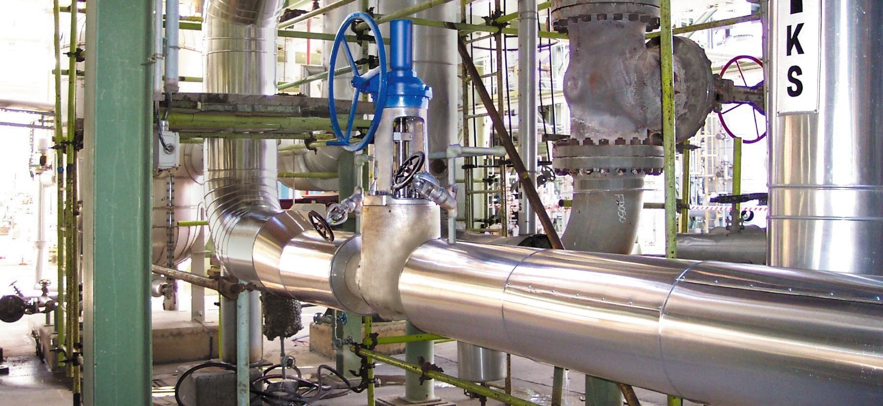 Cadre industriel avec de grands tuyaux métalliques brillants, une vanne à poignée bleue et une armature métallique verte. Diverses jauges et équipements sont visibles, ainsi qu'une étiquette "KS" et une flèche directionnelle sur un tuyau vertical.