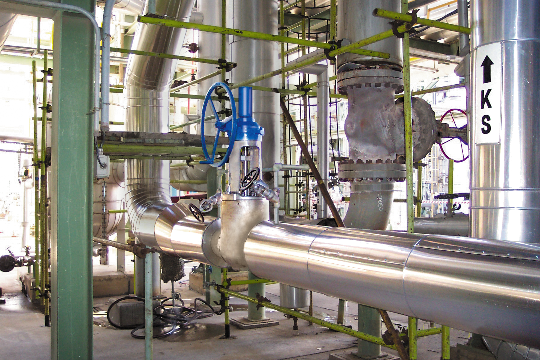 A complex network of shiny metal pipes and valves in an industrial facility, with a blue-handled valve in the center and green structural supports surrounding the equipment.