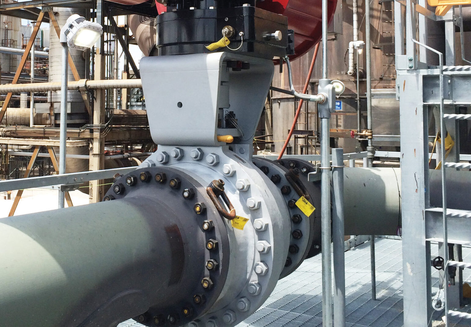Large industrial valve with a red actuator mounted on a pipeline at a refinery or processing plant, surrounded by metal structures and equipment under a blue sky.