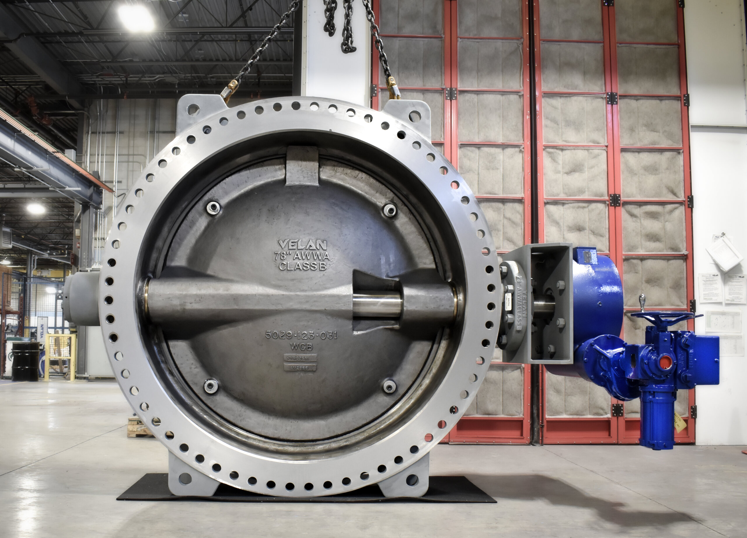 A massive Velan 78-inch AWWA Class B butterfly valve suspended by chains in an industrial facility, with a blue actuator mounted on the side.
