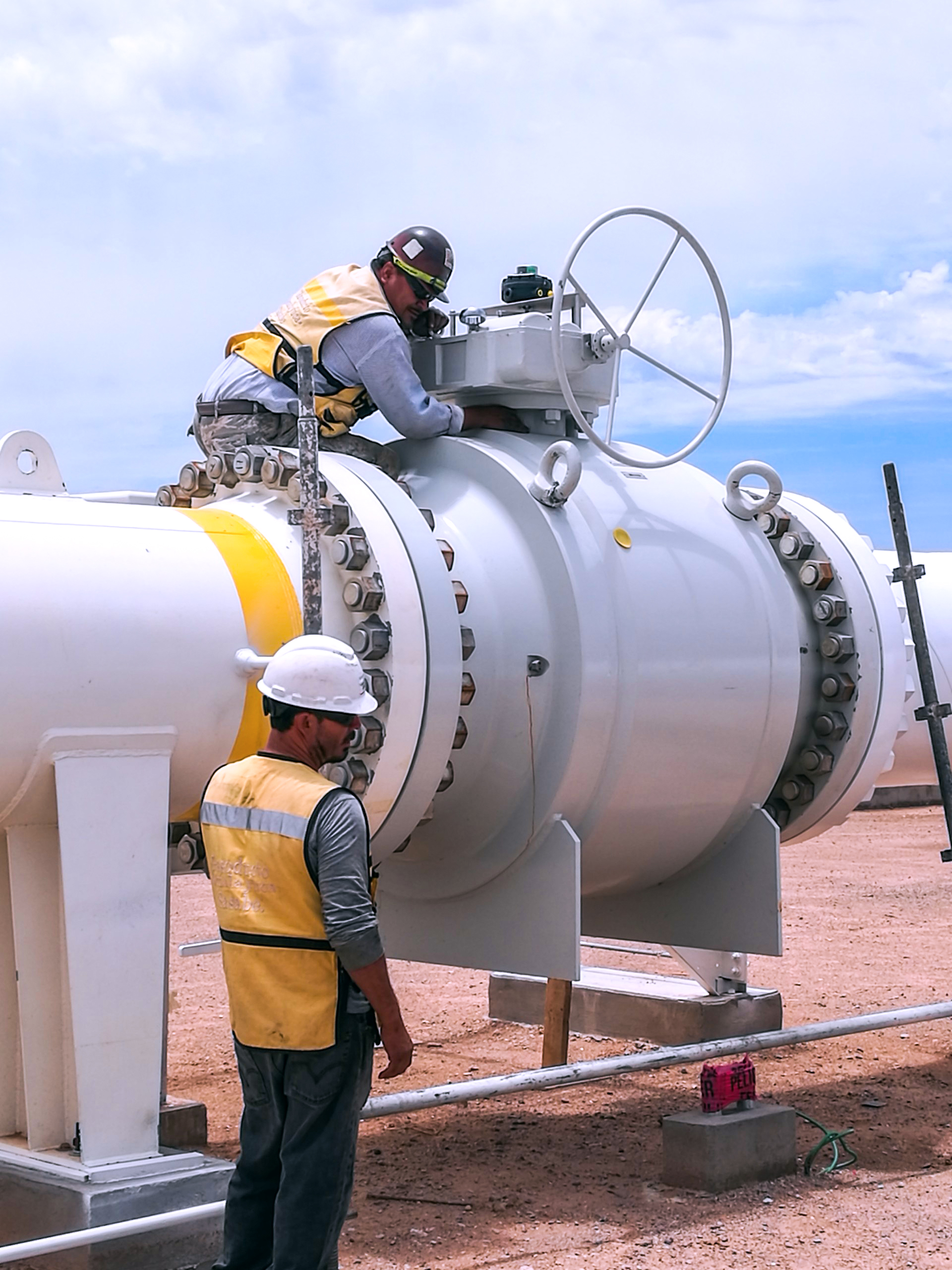 Two workers in safety gear and helmets inspect and work on a large white industrial pipeline valve outdoors on a construction site. One worker is on top of the valve, while the other stands beside it.