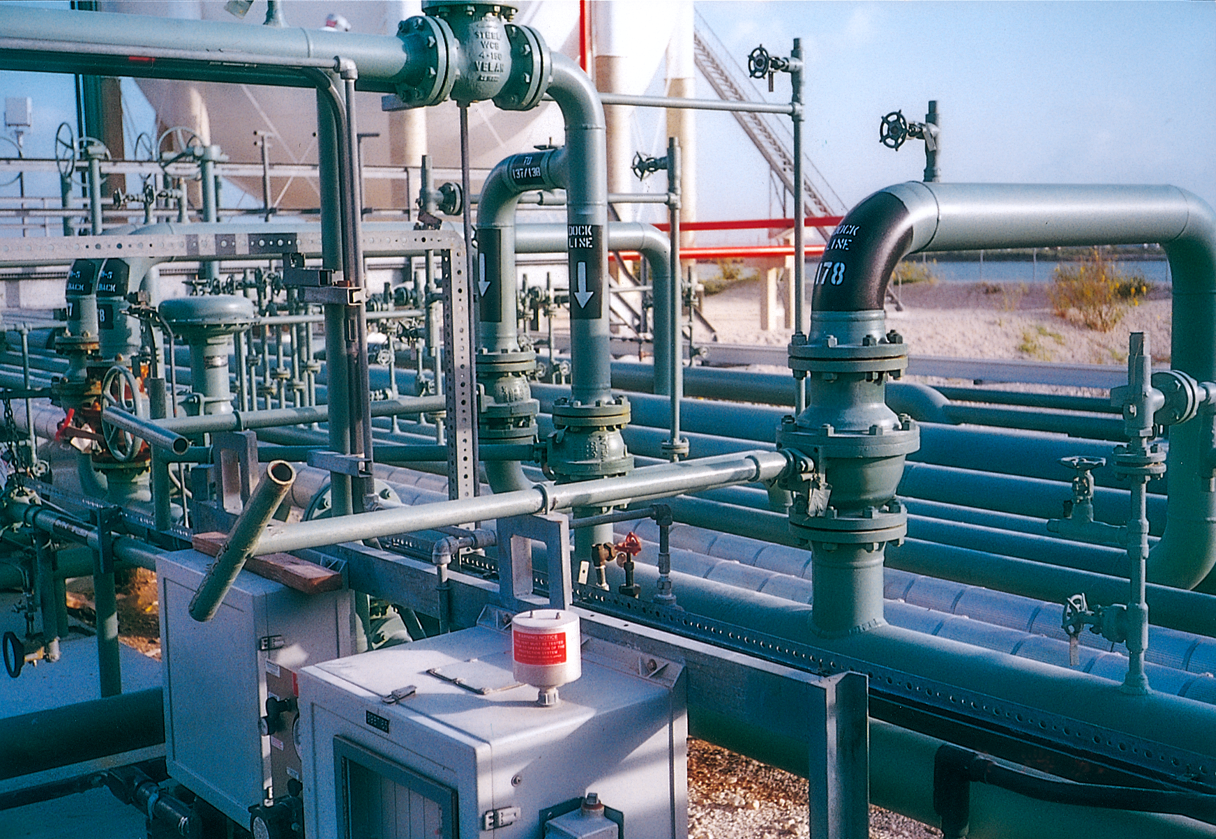 An industrial facility with multiple metal pipes, valves, and gauges, including a red warning light on a control box; the background shows more piping and an outdoor setting under a blue sky.