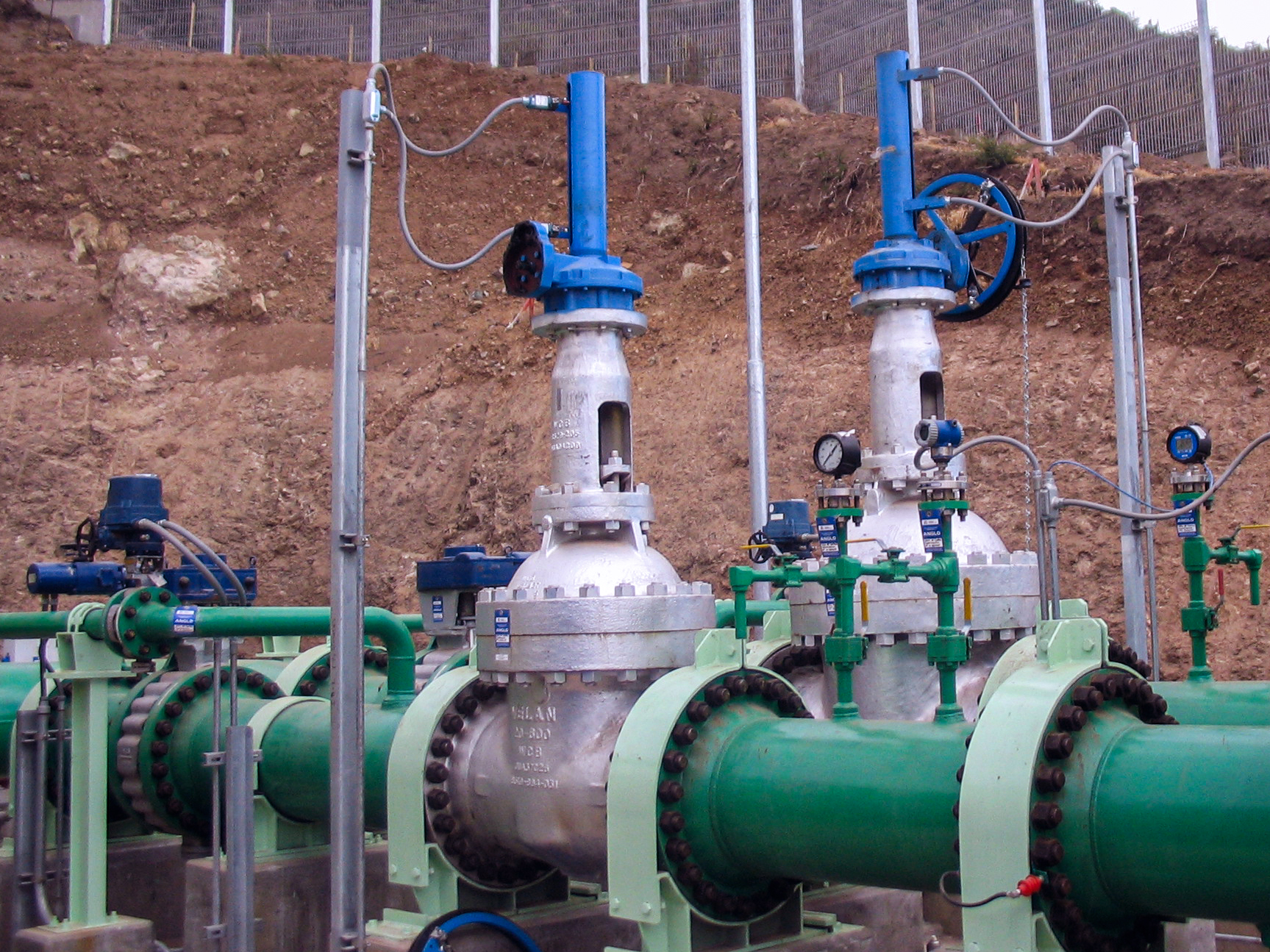 Two large industrial valves with blue actuators are mounted on green pipelines outdoors, set against a dirt embankment and a metal fence in the background. Various gauges, pipes, and wiring are attached to the valves.