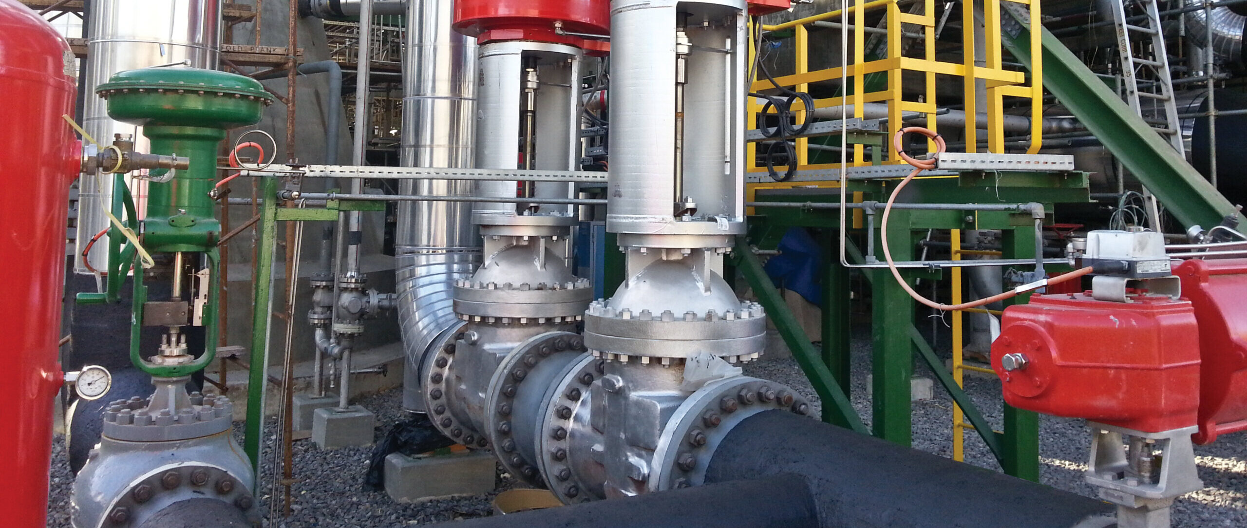 Industrial piping system with large silver valves topped by red actuators, surrounded by metal pipes, green and yellow structural supports, and various mechanical equipment in an outdoor setting.