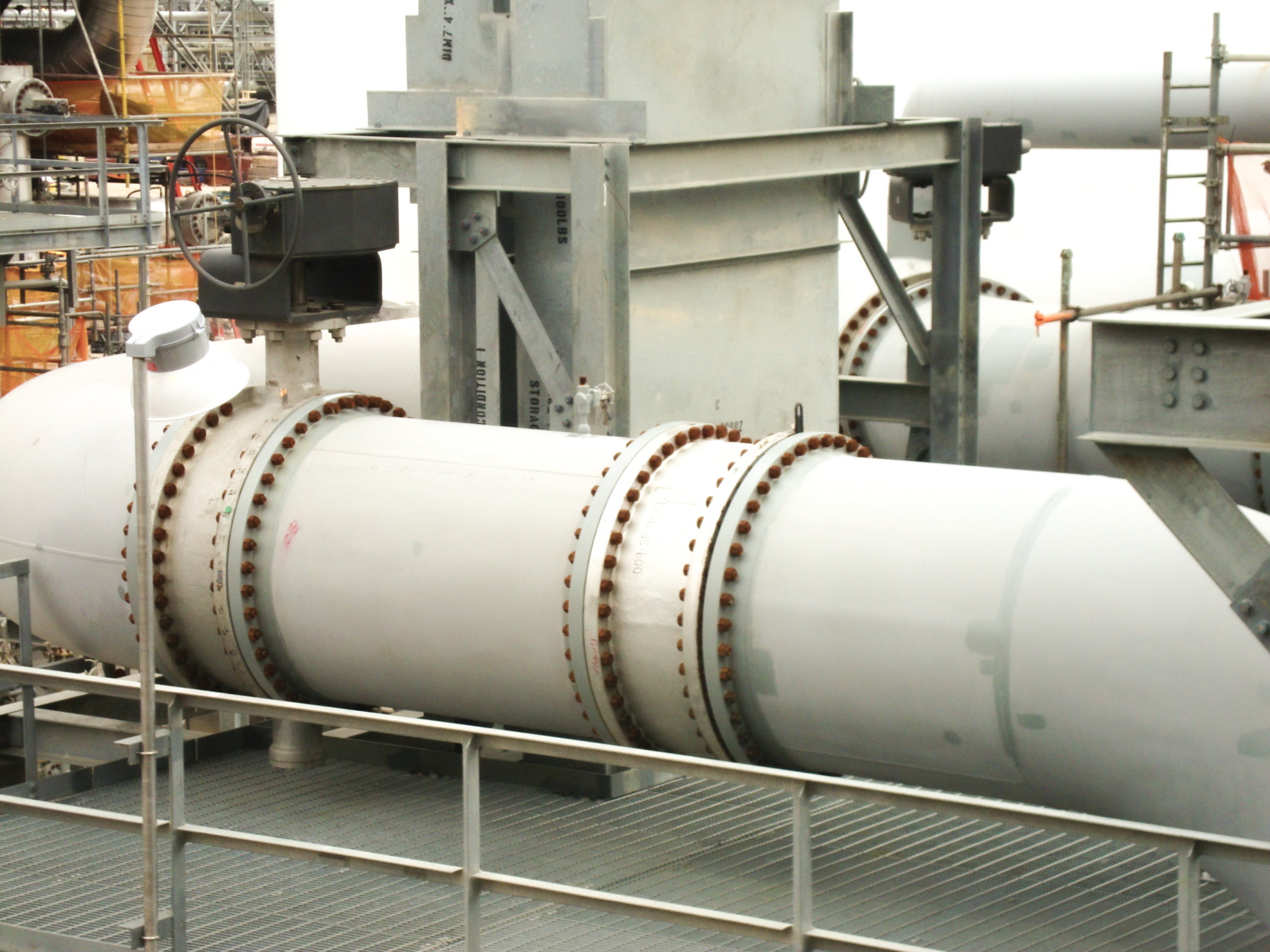 Large industrial pipes with bolted flanges supported by metal structures in a facility, with platforms and railings visible in the foreground. Scaffolding and assorted equipment appear in the background.