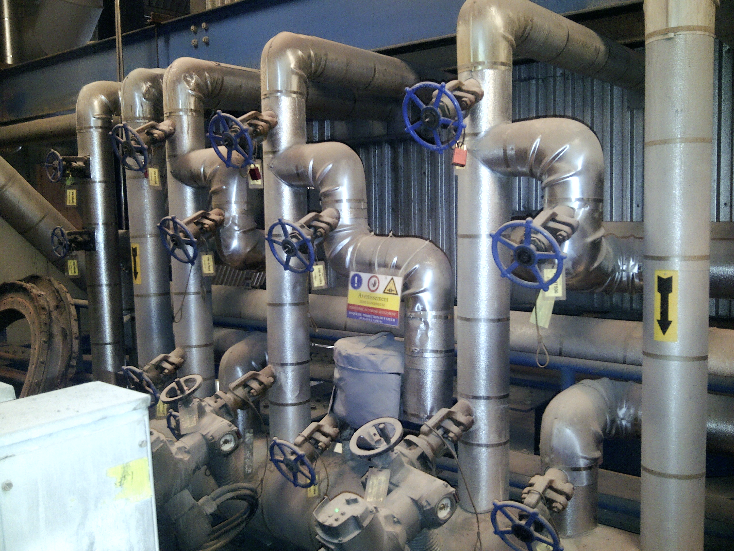 Several insulated industrial pipes with blue-handled valves and warning labels are installed in a facility, with arrows indicating flow direction. The pipes run vertically and horizontally in a complex arrangement.