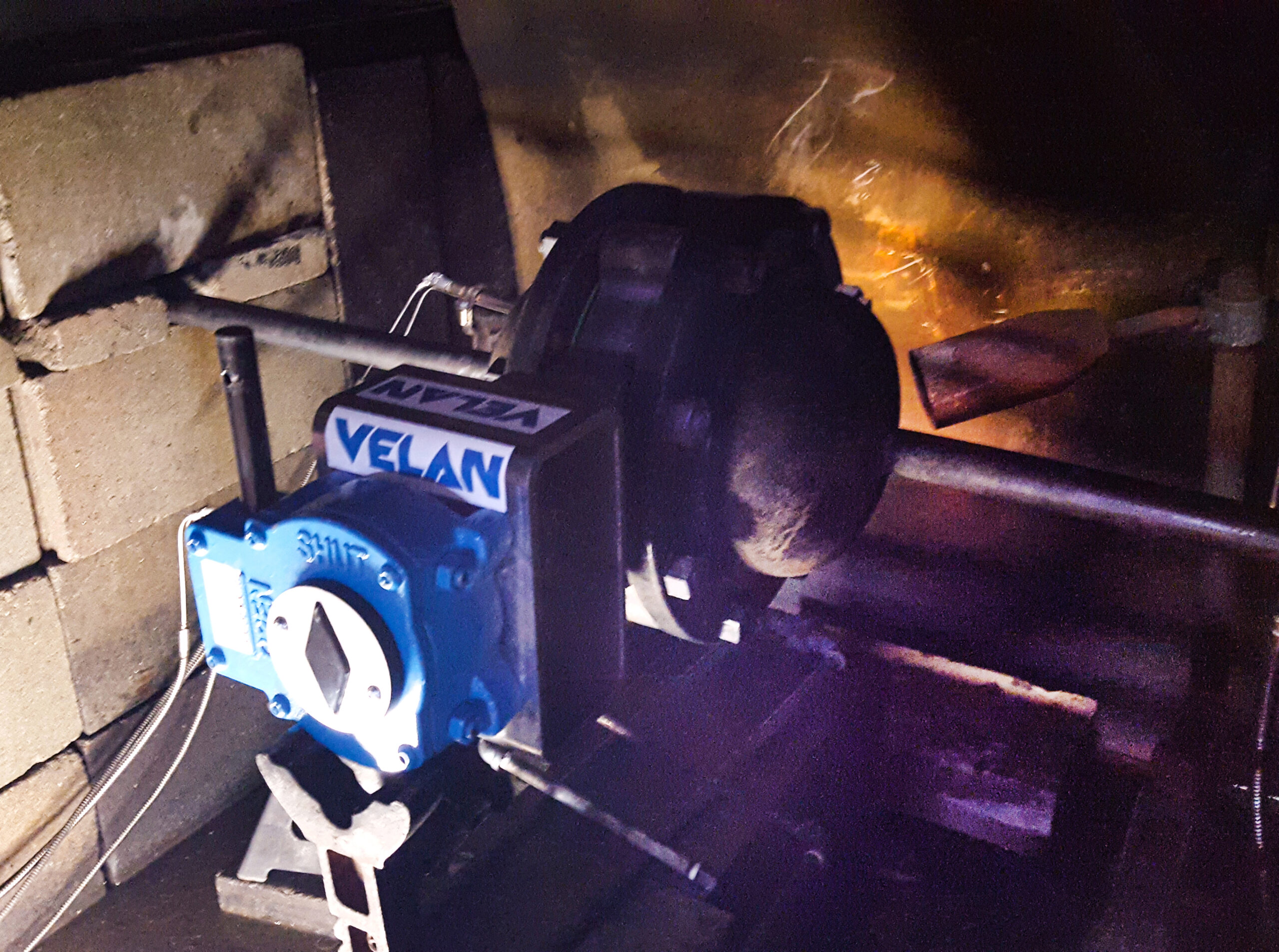 A Velan industrial valve with a blue actuator installed in a dark, confined space near a pipe, with flames or heat visible in the background.