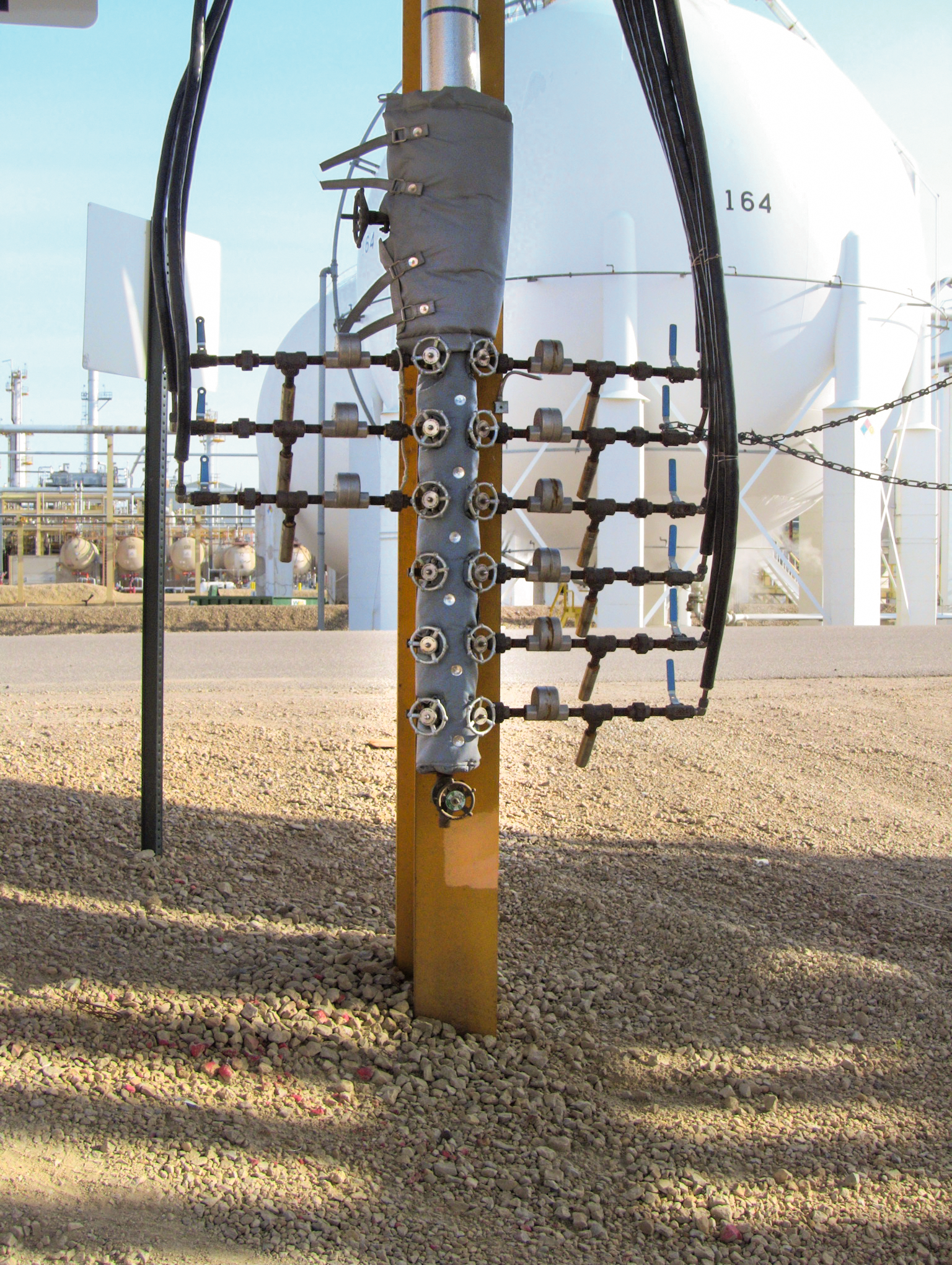 Vertical metal pipe with multiple valves and gauges, some wrapped in insulation, is mounted outdoors on gravel with industrial tanks and equipment visible in the background.