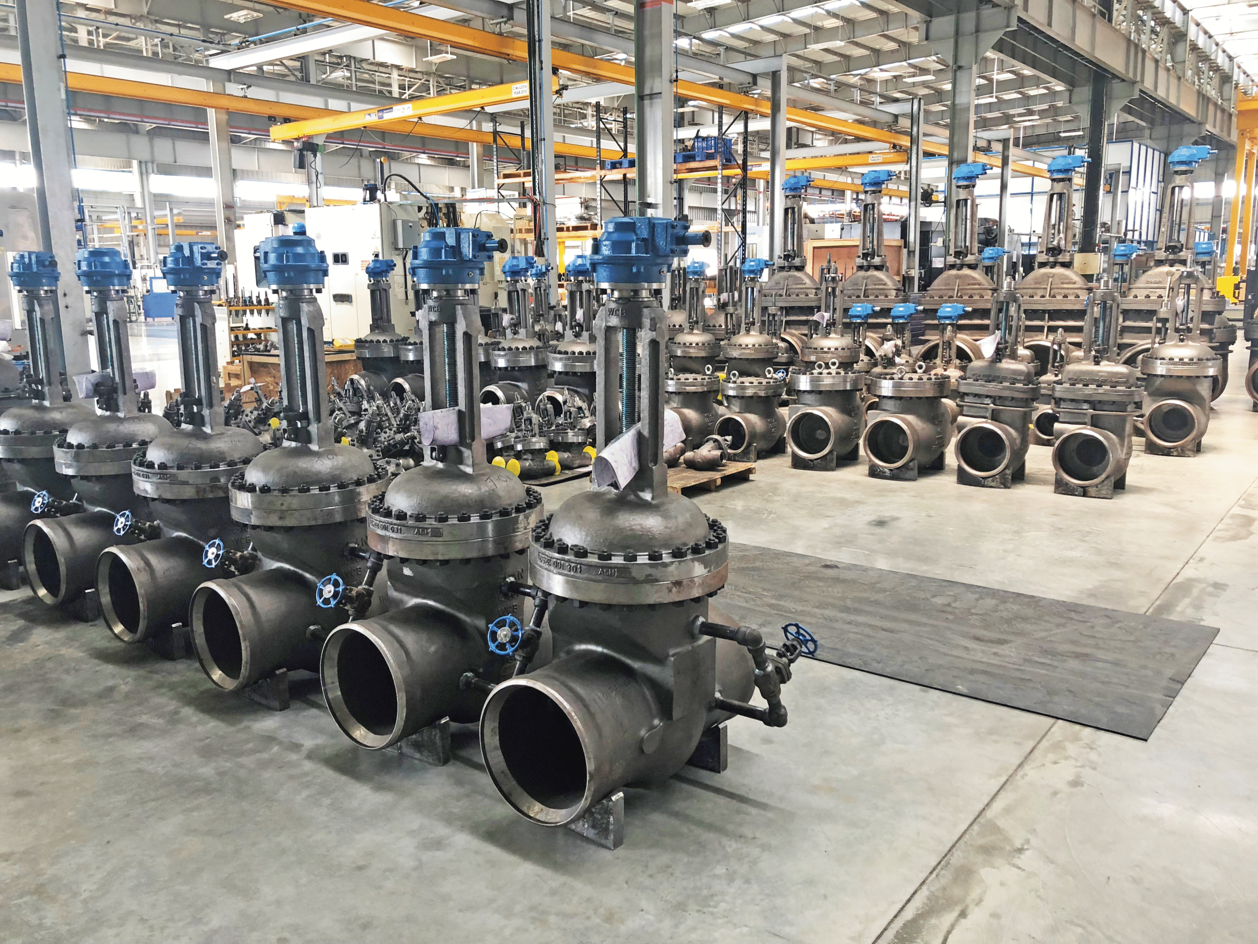 Large industrial metal valves with blue handles are arranged in rows inside a spacious, well-lit factory or warehouse with beams, cranes, and machinery visible in the background.