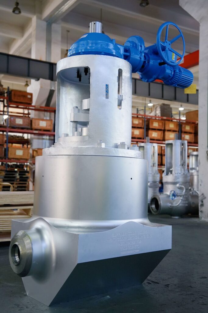 A large industrial metal valve with a silver body and blue actuator sits in a factory workshop, surrounded by shelves with wooden crates and other metal equipment in the background.
