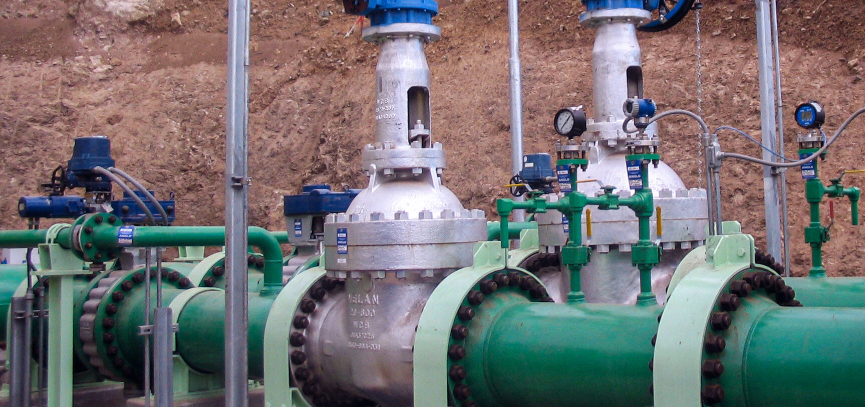De grandes vannes et canalisations industrielles avec des tuyaux verts et des raccords métalliques argentés sont installées à l'extérieur, contre un talus de terre. Des actionneurs bleus, des manomètres et des équipements de contrôle sont montés sur les tuyaux.
