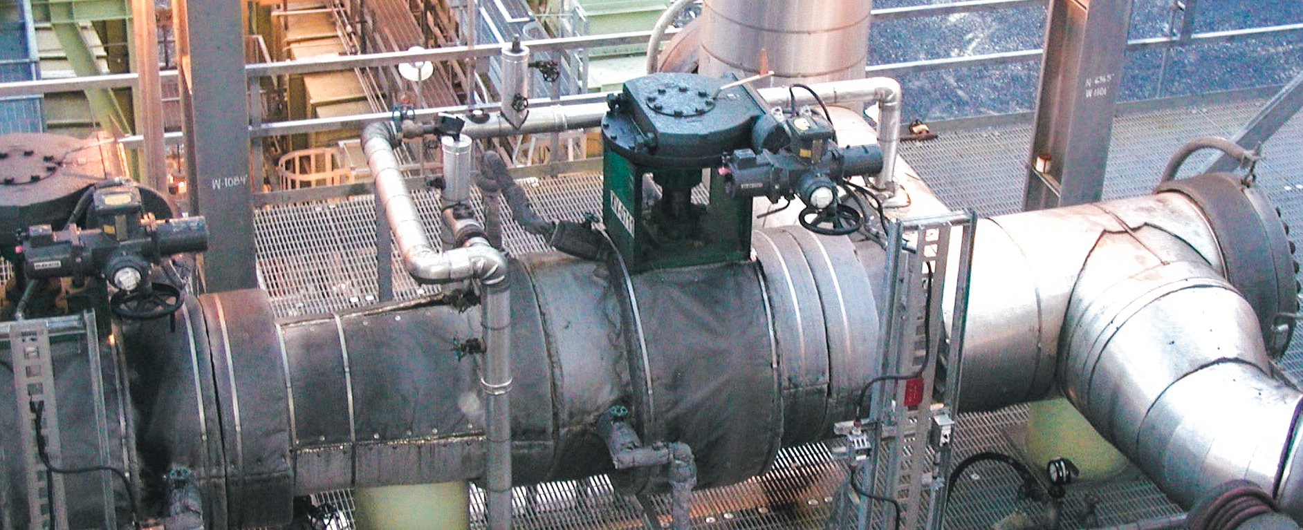Large industrial pipes and valves in a processing plant, with various metal walkways, handrails, and overhead lights. The setting appears to be a facility for water treatment, chemical processing, or energy production.