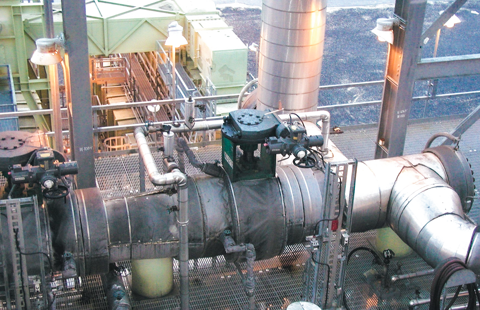 Gros tuyaux et valves industriels à l'intérieur d'une usine de transformation, entourés de structures métalliques, de lampes et de passerelles, avec divers équipements et machines visibles à l'arrière-plan.