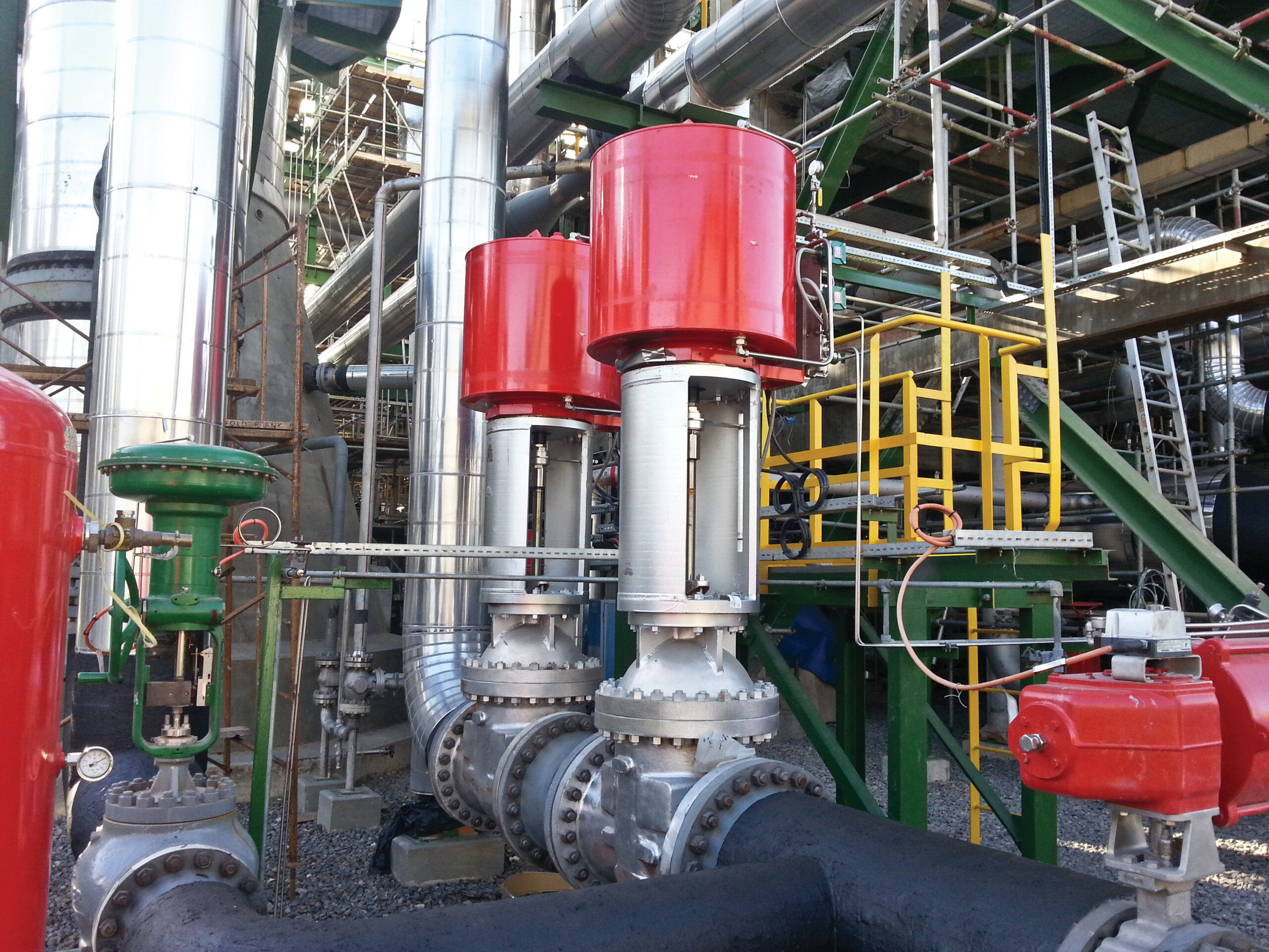 De grandes vannes industrielles avec des actionneurs rouges sont installées sur des tuyaux métalliques dans une usine de transformation. L'arrière-plan est constitué d'échafaudages métalliques, d'escaliers et de divers éléments de machines.