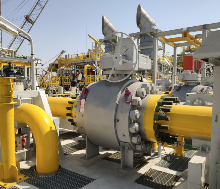 Un grand ensemble de vannes industrielles avec des tuyaux jaunes est situé dans une installation extérieure, entouré de diverses machines et structures métalliques sous un ciel clair.