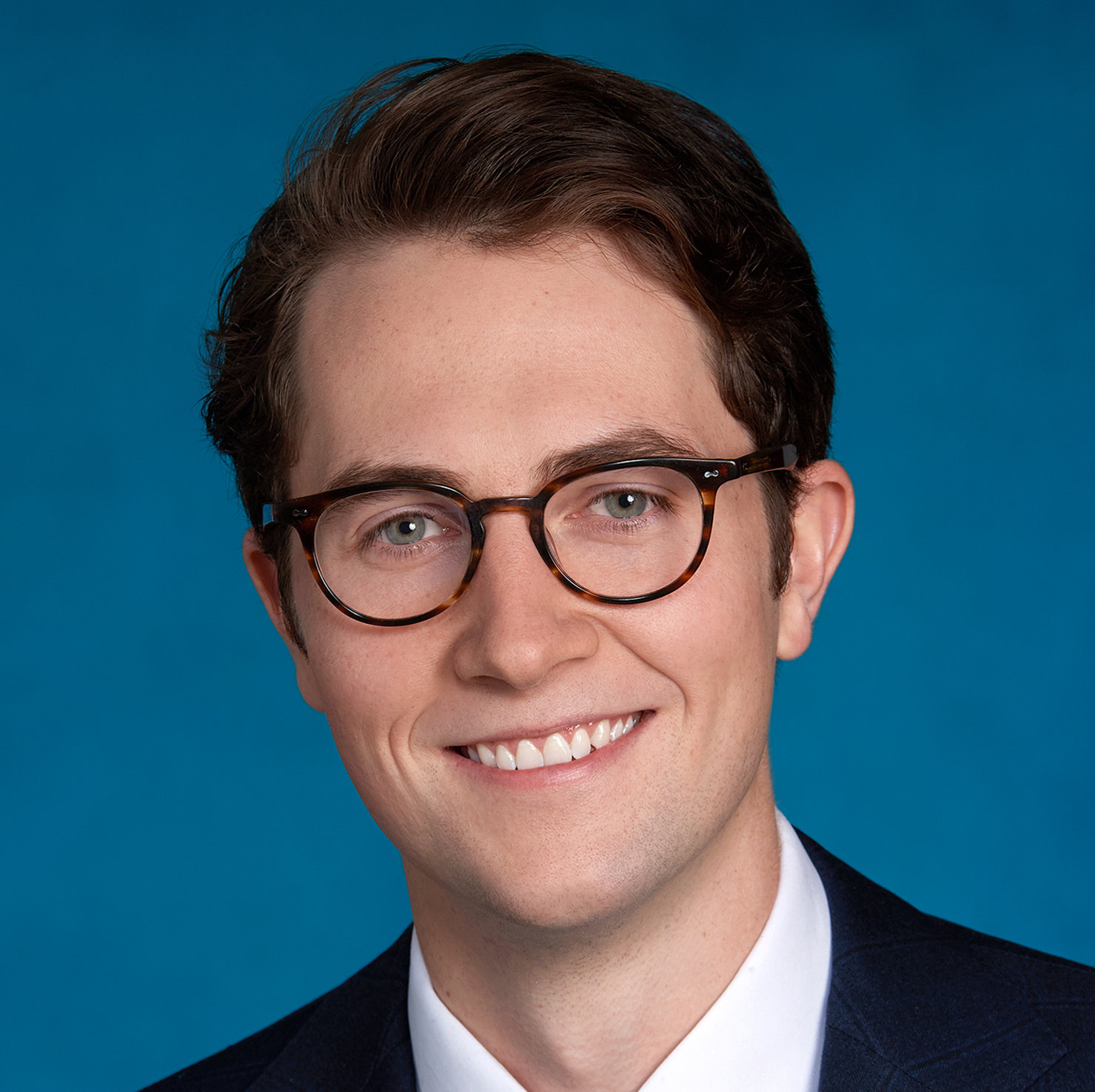Un jeune homme aux cheveux bruns courts et portant des lunettes, vêtu d'un costume sombre et d'une chemise blanche, souriant, posait devant un fond bleu uni.
