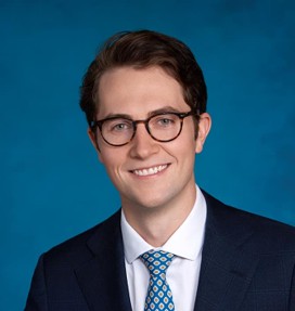 Un jeune homme aux cheveux bruns courts et portant des lunettes, vêtu d'un costume sombre, d'une chemise blanche et d'une cravate bleue à motifs, sourit devant un fond bleu uni.