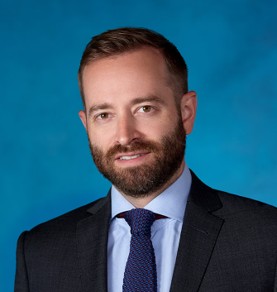 Un homme aux cheveux bruns courts et à la barbe, vêtu d'un costume sombre, d'une chemise bleu clair et d'une cravate bleu marine texturée, pose devant un fond bleu uni.