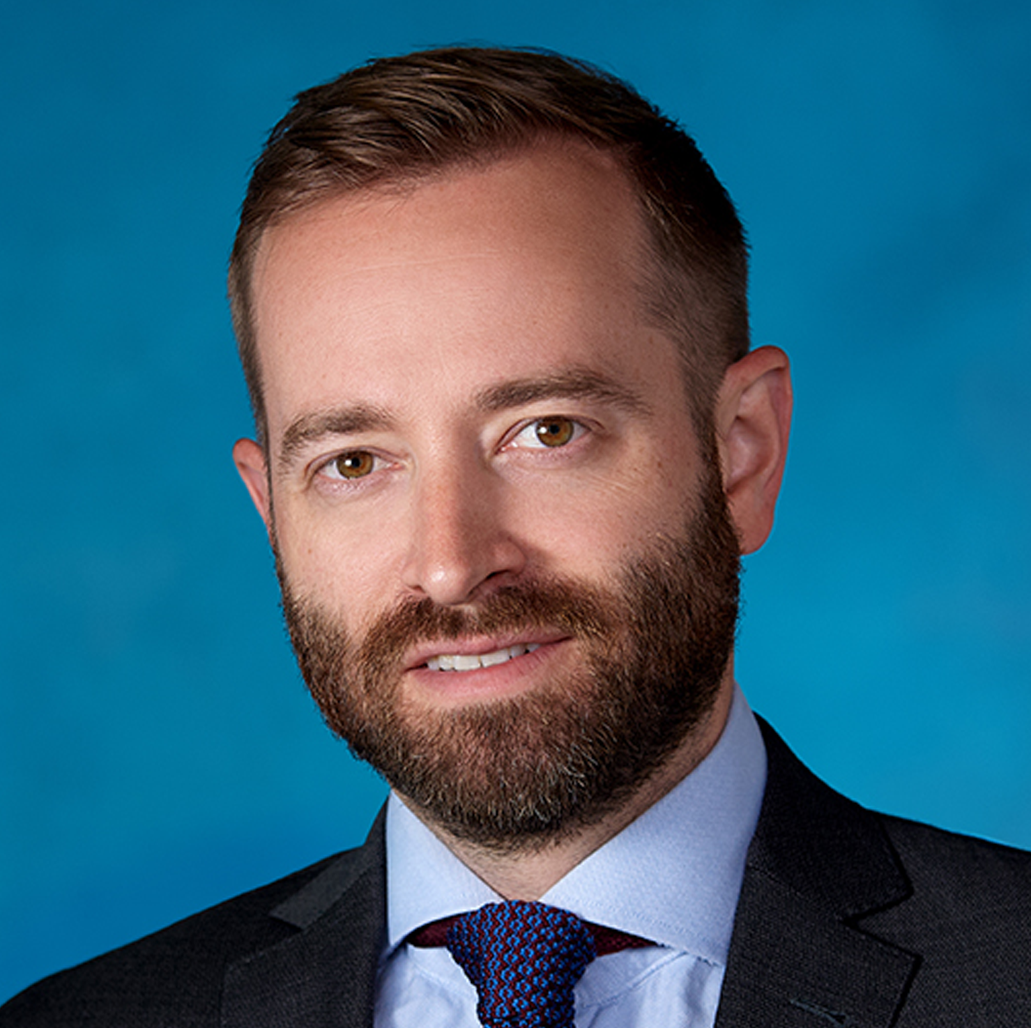 Un homme aux cheveux bruns courts et à la barbe, vêtu d'un costume sombre, d'une chemise bleu clair et d'une cravate à motifs, pose devant un fond bleu uni.