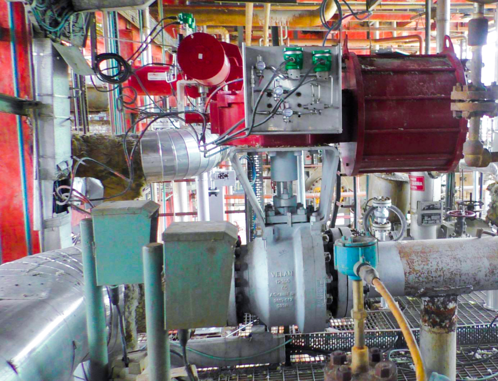 Industrial valve system with attached actuators, control boxes, and wiring; various pipes and metal structures surround the equipment inside a plant facility. Red and green actuator components are prominently visible.