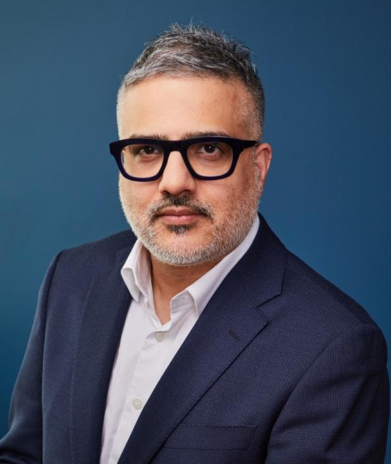 A man with short gray hair and a beard, wearing black glasses, a white shirt, and a dark blue suit jacket, poses against a plain blue background.