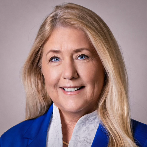 A woman with long blonde hair and blue eyes smiles at the camera. She is wearing a blue blazer over a white blouse, and the background is plain and light-colored.