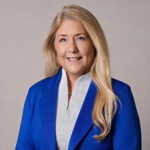 Une femme aux longs cheveux blonds sourit à la caméra. Elle porte un blazer bleu roi par-dessus un chemisier blanc et se tient debout devant un fond uni gris clair.