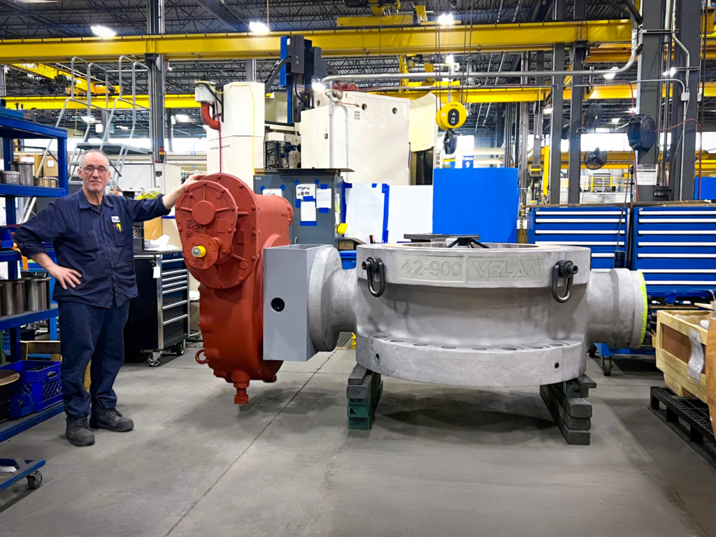 A man in work clothes stands next to a large industrial valve inside a factory, surrounded by machines and blue tool cabinets. The valve is silver and red, marked "VELAN 42-900.