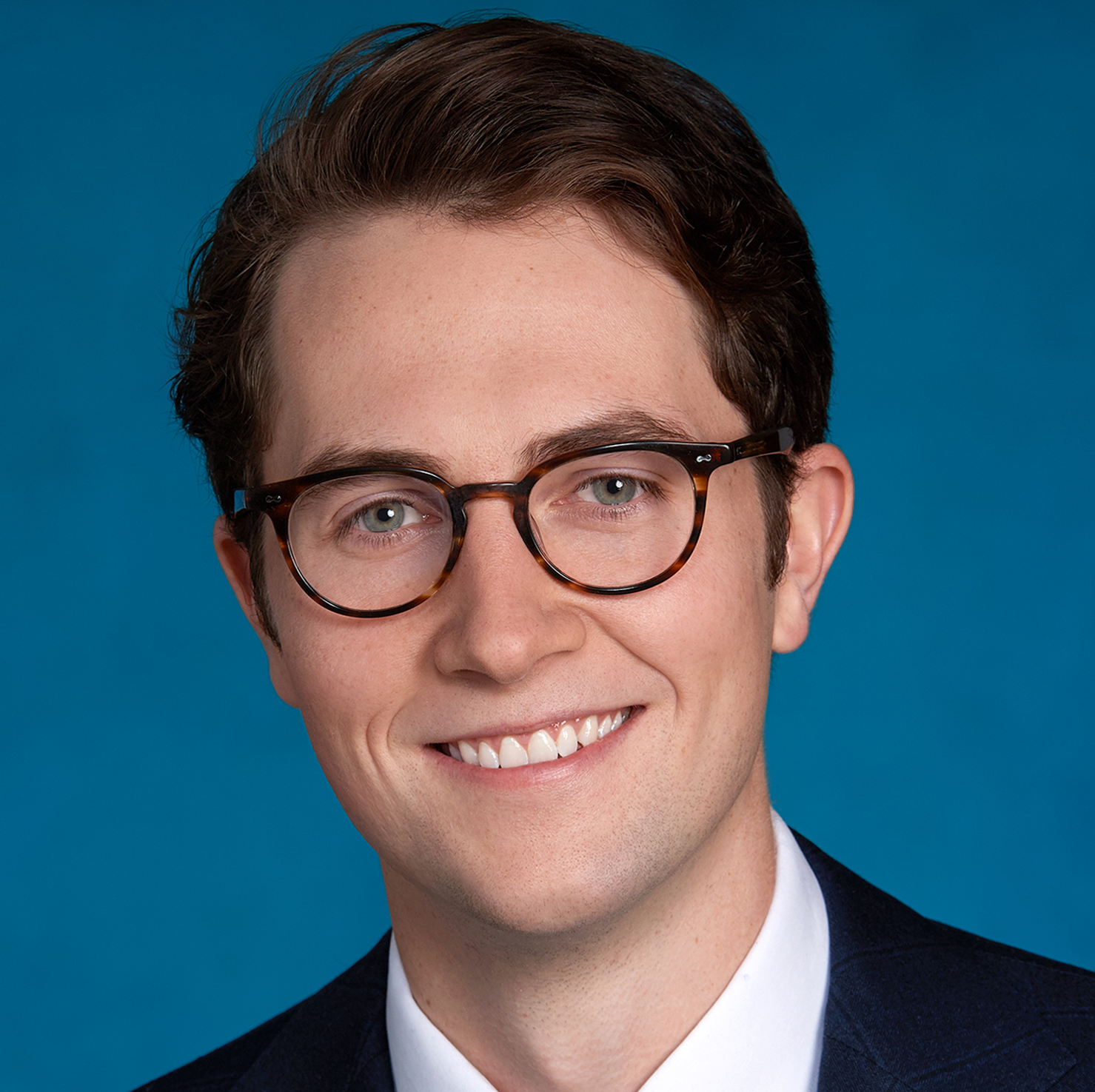 A young man with short brown hair and glasses, wearing a dark suit jacket, white shirt, and tie, smiles in front of a solid blue background.