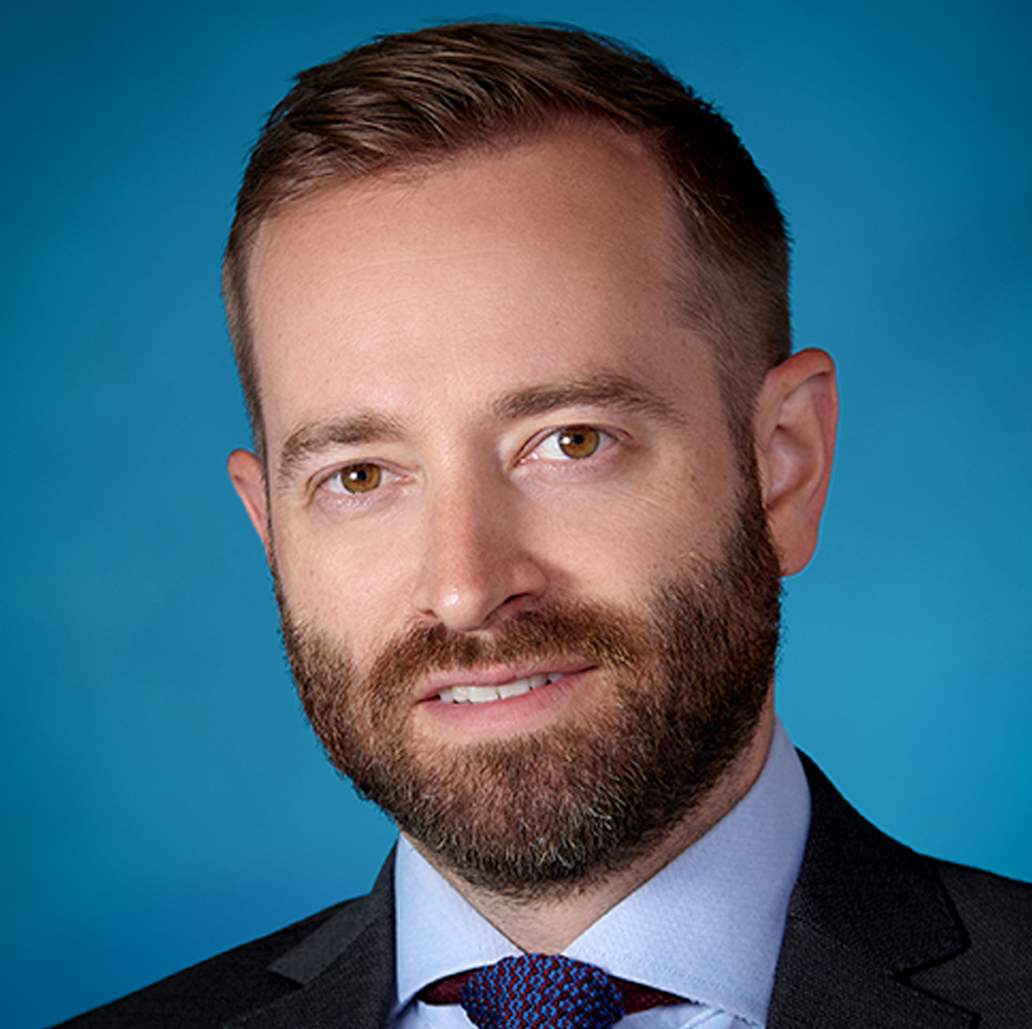 A man with short brown hair and a trimmed beard, wearing a gray suit, light blue shirt, and patterned tie, poses in front of a blue background.